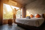 Portrait of tourist woman raised her hands and standing nearly window, looking to beautiful view with her luggage in hotel bedroom after check-in.