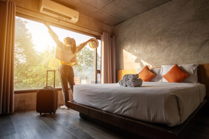 Portrait of tourist woman raised her hands and standing nearly window, looking to beautiful view with her luggage in hotel bedroom after check-in. Hotel guest arriving in guestroom.