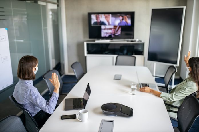 Two people sit at a conference table waving at three people attending virtually visible on a screen at a hybrid meeting